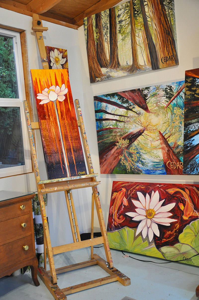 Artwork in Cedar Lee studio. On easel: "Free Spirit." Background, left-right, top-bottom: "Lotus Study 2," "Forest Nymph," "Dance of the Redwoods," "Koi Circles"