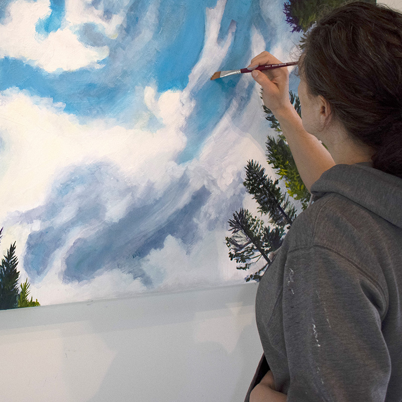 Artist Cedar Lee working on her painting of  clouds floating in a blue sky over evergreen trees.