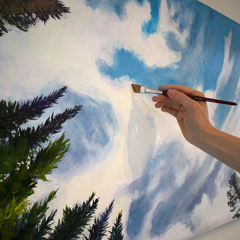 Artist Cedar Lee working on painting of clouds floating in a blue sky over evergreen trees.