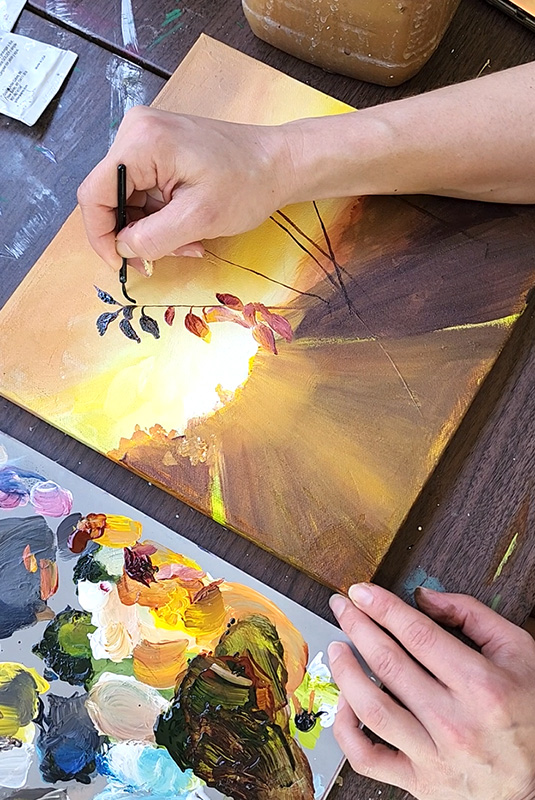 Cedar Lee painting a work in progress of a sunset painting on a small canvas, with a paint palette full of mixed paint colors.